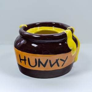 Può includere: Un vasetto di miele in ceramica con esterno marrone e interno giallo. Il vaso ha una striscia gialla con la scritta "HUNNY" in lettere nere. Il miele giallo cola lungo il lato.