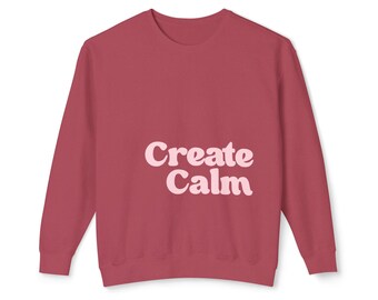 May include: A dark red crewneck sweatshirt featuring the text "Create Calm" printed in a light pink, rounded, retro-style font across the center of the chest. The garment has long sleeves, a ribbed neckline, and ribbed cuffs at the wrists and waist.