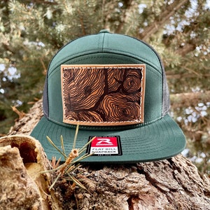 May include: A green flat bill snapback hat with a black mesh back. The front features a brown leather patch with a tree ring design. The hat has a white stitched brim and a tag that reads "Flat Bill Snapback".