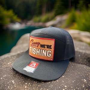 May include: A black flat bill snapback hat with a mesh back. The hat features a brown leather patch with the words "Sorry We're Fishing" in white and red lettering. A tag with the brand name is attached to the brim.