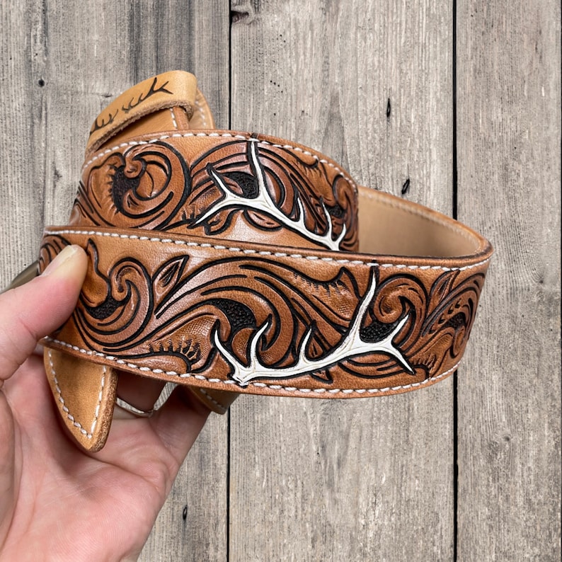 May include: A brown leather belt with a detailed carved floral pattern and white antler accents. The belt is stitched with white thread and has a natural leather finish. The belt is approximately 1.5 inches wide.