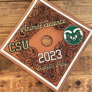 May include: A brown leather graduation cap with the words "Animal Science" and "CSU" in gold and green. The year "2023" and the name "Isabella May" are also featured. The cap has a white border and decorative floral tooling.