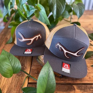 May include: Two gray flat bill snapback hats with tan mesh backs. Each hat features a white and brown antler design on the front. The hats are displayed on a wooden surface with green foliage in the background.