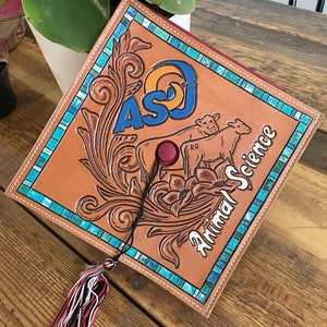 May include: A brown leather graduation cap with the text "ASJ" in blue and orange, and "Animal Science" in white. The cap features a floral design and two cow illustrations. A tassel with red, black, and white strands hangs from the cap.