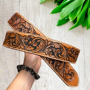 May include: A brown leather belt with intricate floral and scrollwork carving in black. The belt has a light brown border and white stitching. The belt is held up against a wooden background with green leaves.