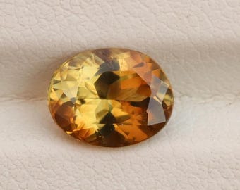 Könnte beinhalten: Ein facettierter, oval geformter Edelstein mit einer Mischung aus Gelb-, Orange- und Brauntönen. Der Edelstein ist vor einem neutralen, strukturierten Hintergrund platziert, der seinen Brillantschliff und seine Farbvariationen hervorhebt. Geeignet für die Schmuckherstellung.