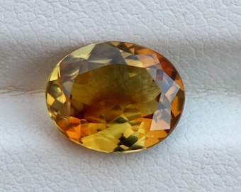 Könnte beinhalten: Ein facettierter, ovaler Edelstein mit warmen Farben. Der Edelstein weist Gelb-, Orange- und Brauntöne auf und hat eine helle, reflektierende Oberfläche. Der Edelstein liegt vor einem weißen Hintergrund.