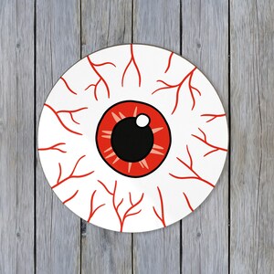 Puede incluir: Un posavasos redondo blanco con un diseño de ojo de dibujos animados. El ojo tiene un iris rojo, una pupila negra y venas rojas que irradian hacia afuera. Perfecto para Halloween o decoración espeluznante.