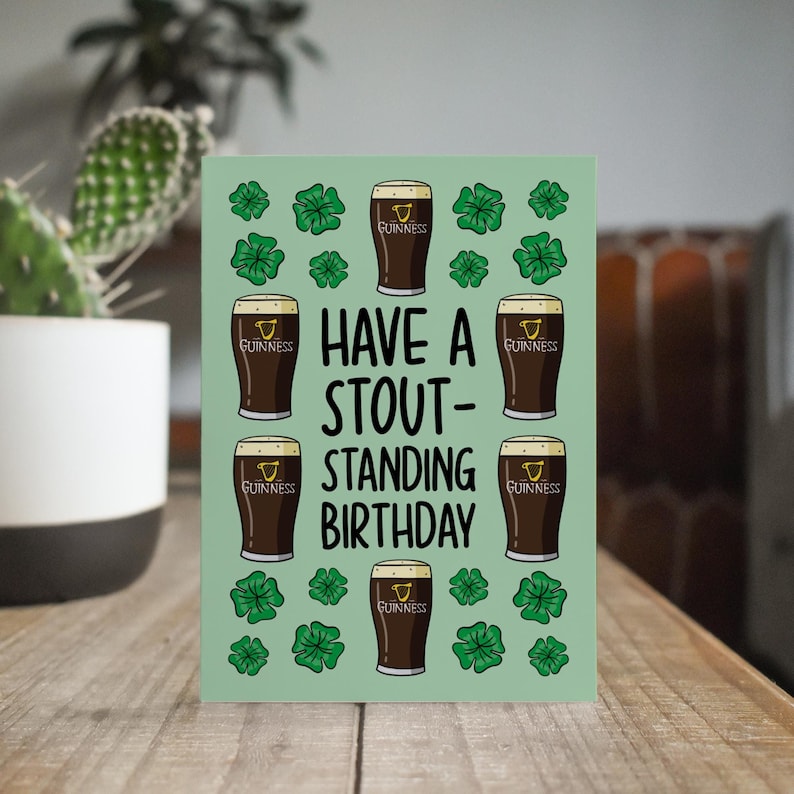 Op de afbeelding: Een groene kaart met een verjaardagsgroet die "Have a Stout-Standing Birthday" zegt. De kaart is versierd met klavertjesvier en illustraties van glazen Guinness-bier.