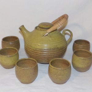 May include: A set of six green and brown ceramic teacups and a teapot with a woven handle. The teapot has a round body and a lid. The cups are all the same size and shape.