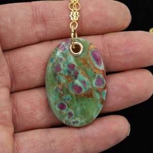 May include: A gold chain necklace with a green and pink oval pendant. The pendant is made of a natural stone with a speckled pattern.