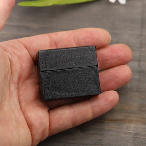 May include: A small black square box with a textured surface. The box is closed and is being held in a person's hand.
