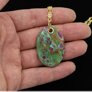 May include: A gold chain necklace with a green and pink oval pendant. The pendant is made of a natural stone with a speckled pattern.