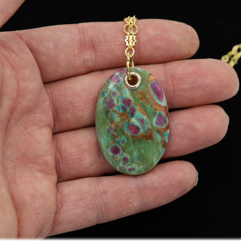 May include: A gold chain necklace with a green and pink oval pendant. The pendant is made of a natural stone with a speckled pattern.