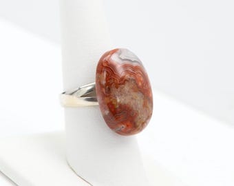 May include: A silver ring with a large, oval-shaped agate gemstone. The agate is a mix of red, brown, white, and cream colors with a swirling pattern.