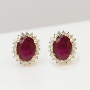 May include: A pair of gold earrings featuring oval ruby gemstones set in a halo of smaller diamonds.