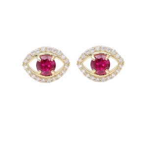 May include: A pair of gold-toned stud earrings, each designed as an eye. The center of each eye features a round, vibrant red gemstone, surrounded by a halo of small, clear gemstones. The eye-shaped frame is also adorned with small, clear gemstones.