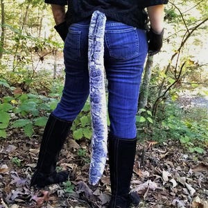 May include: A long, plush, faux fur tail in shades of gray and white, attached to the back of blue jeans. The tail hangs down, contrasting with the dark clothing and boots. The scene is set outdoors.