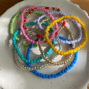 May include: A collection of colourful beaded bracelets in shades of pink, yellow, green, blue, and silver. The bracelets are made of small, round beads and are stacked on top of each other.