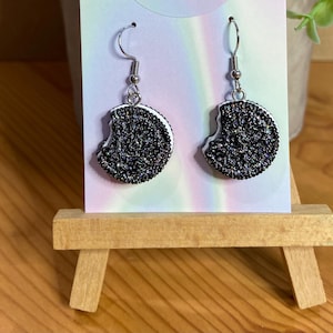 May include: A pair of silver dangle earrings with a black and white cookie charm. The cookie charm is a half-eaten cookie with a white filling and black sprinkles.