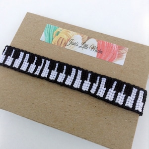 May include: A black and white friendship bracelet with a piano key design. The bracelet is on a brown paper background with a label that says "Jasie's Little Mocha".