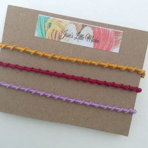 Può includere: Tre fili di cordoncino ritorto in giallo senape, rosso borgogna e lavanda. I cordoncini sono esposti su un cartoncino marrone con un'etichetta che recita "Jassie's Little Mocha".