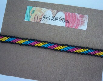 Può includere: Un braccialetto dell'amicizia nero, giallo, rosa e blu con un motivo diagonale. Il braccialetto è su uno sfondo marrone con un'etichetta che recita "Josie's Little Mocha".
