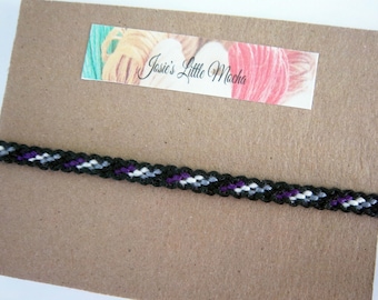 May include: A black, white, gray, and purple friendship bracelet with a woven pattern. The bracelet is on a brown background with a label that says "Josie's Little Mocha".