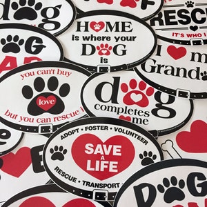 May include: A collection of oval-shaped stickers with various dog-themed designs. The stickers feature black and white paw prints, red hearts, and white text on a white background. Some of the stickers have black text on a white background. The text on the stickers includes phrases like "You had me at woof", "Home is where your dog is", "Dog completes me", and "Save a life".
