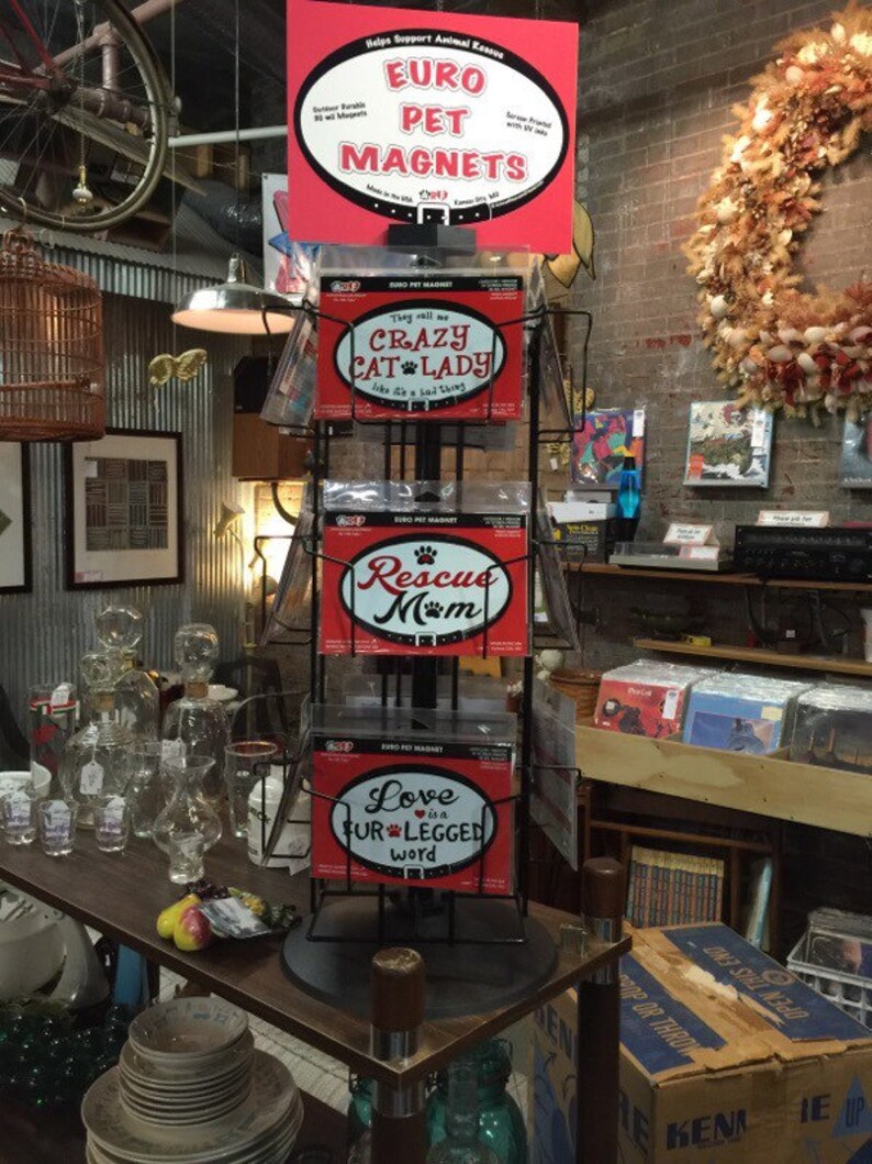 May include: A rotating display stand with four Euro Pet Magnets featuring colorful designs. The magnets feature the text "They will all be Crazy Cat Lady", "Rescue Mom", "Love is a Fur-legged Word", and "Euro Pet Magnets".