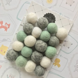 May include: A garland of felt balls in white, grey, and light green. The balls are arranged in a random pattern and are strung together with a clear thread. The garland is on a white background with a black and white pattern.