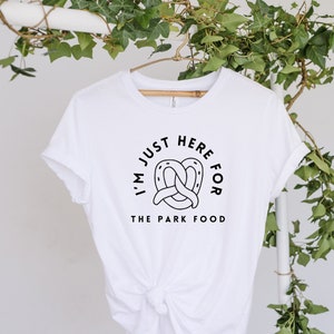 May include: White t-shirt featuring the text "I'M JUST HERE FOR THE PARK FOOD" in a black circular design, with a pretzel graphic. The shirt is displayed on a wooden hanger, set against a backdrop of green foliage.