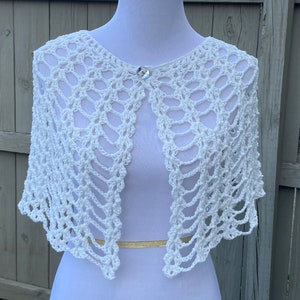 May include: A white, open-weave crochet shawl with a delicate pattern. The shawl is fastened with a sparkling, round button. The garment is displayed on a white mannequin, showcasing its open front design. A gold-colored band is visible at the waist.