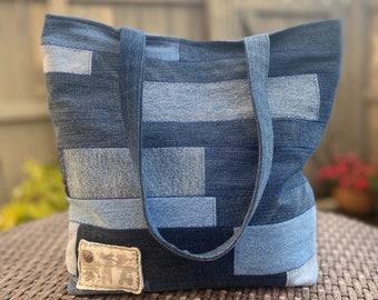 May include: A blue denim tote bag made from recycled jeans. The bag has a patchwork design with different shades of blue denim. The bag has two long straps and a small leather patch with the word "Silver" on it.