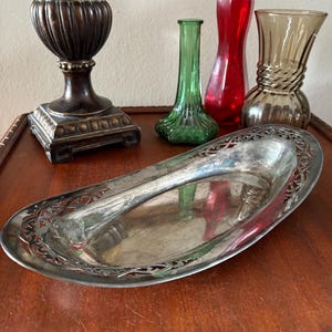 May include: A silver-colored, oval-shaped serving tray with an ornate, openwork border. The tray sits on a wooden surface, alongside decorative glass vases in green, red, and amber hues, and a bronze-colored decorative lamp.