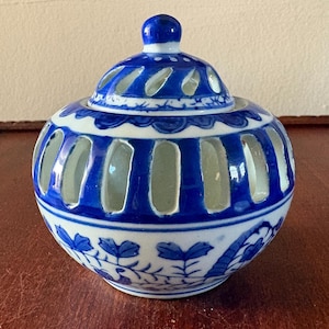 May include: A blue and white ceramic jar with a lid. The jar has a round shape with vertical cut-out openings. The lid has a small knob and cut-out designs. Floral patterns decorate the base of the jar.