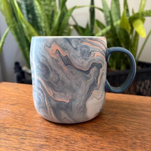 May include: A ceramic mug with a marbled design in shades of gray, peach, and white. The mug has a dark blue handle and a rounded shape. The mug is sitting on a wooden surface.