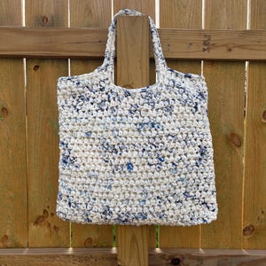 May include: A handmade tote bag with a white and blue speckled pattern. The bag is made of a thick, woven material and has two handles. The bag is hanging on a wooden fence.