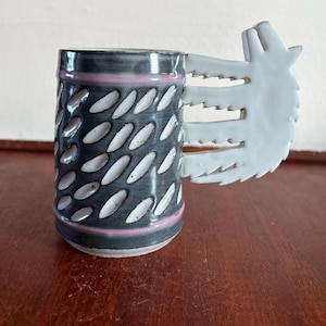 May include: A ceramic mug with a unique design. The mug features a dark gray body with white oval accents and a light pink band. The handle is shaped like a wolf's head. The mug is on a wooden surface.
