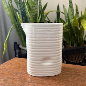 May include: A white ceramic pitcher with a ribbed design. The pitcher has a spout and a cylindrical shape. The background includes green plants and a wooden surface.