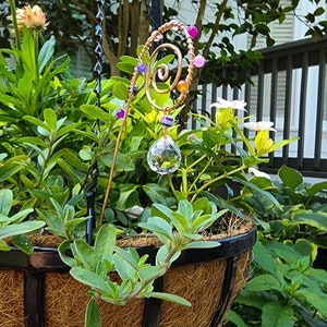 Handmade plant stake suncatcher for garden decor, crafted with 16-gauge copper wire in a spiral design. The stake is decorated with colorful glass beads and features a millefiori glass bead hanging above a 20mm K9 crystal ball. The crystal sparkles in the sunlight, creating rainbow light effects among the plants.