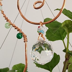 Close-up of a handmade plant stake suncatcher for garden decor, crafted with 16-gauge copper wire in a spiral design. The stake is adorned with colorful glass beads and features a millefiori glass bead hanging above a 20mm K9 crystal ball. The crystal sparkles in the sunlight, scattering rainbow light effects.