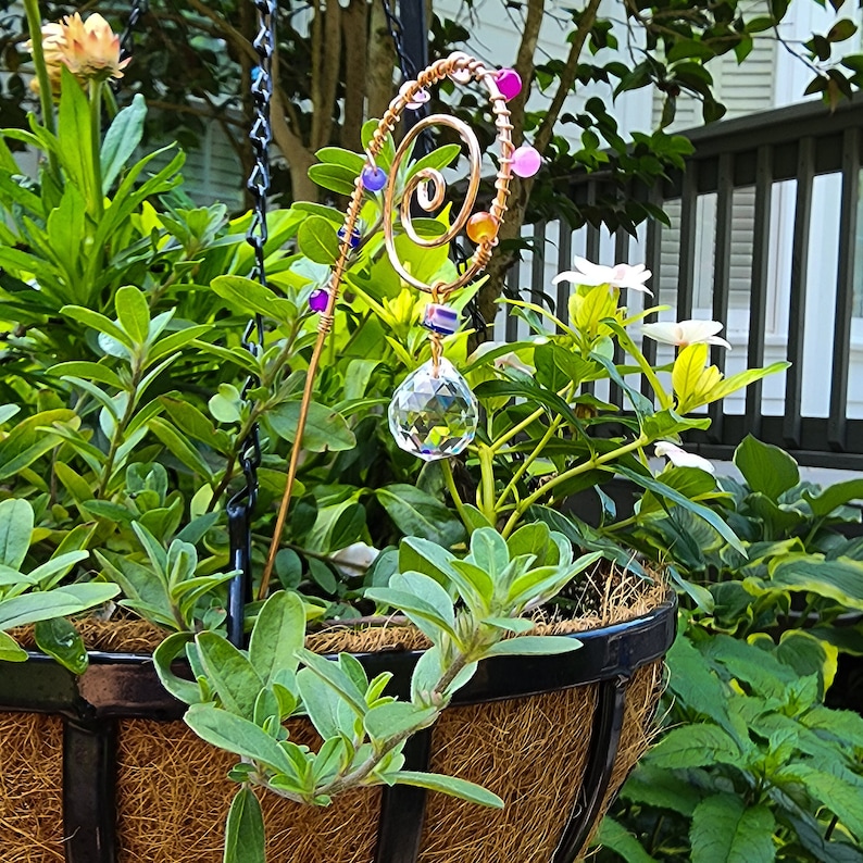 Handmade plant stake suncatcher for garden decor, crafted with 16-gauge copper wire in a spiral design. The stake is decorated with colorful glass beads and features a millefiori glass bead hanging above a 20mm K9 crystal ball. The crystal sparkles in the sunlight, creating rainbow light effects among the plants.