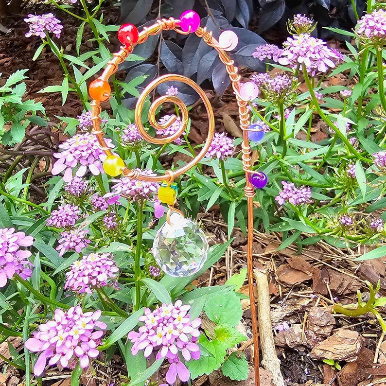 Handmade plant stake suncatcher for garden decor, crafted with 16-gauge copper wire in a spiral design. The stake is decorated with colorful glass beads and features a millefiori glass bead hanging above a 20mm K9 crystal ball. Displayed outdoors among vibrant red flowers and green foliage, the crystal sparkles in the sunlight with rainbow light effects.