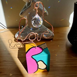 May include: A copper wire sculpture spelling "love" with a clear crystal prism hanging from the top. Below is a small, colorful wooden block painted with abstract shapes in pink, yellow, and teal. The items are on a wooden surface.