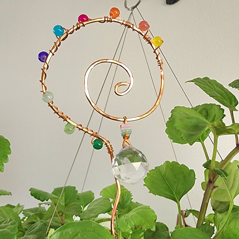 Close-up of a handmade plant stake suncatcher for garden decor, crafted with 16-gauge copper wire in a spiral design. The stake is adorned with colorful glass beads and features a millefiori glass bead hanging above a 20mm K9 crystal ball. The crystal sparkles in the sunlight, scattering rainbow light effects.