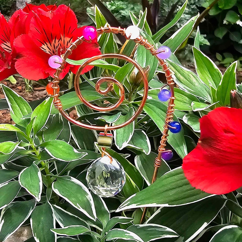 "Handmade plant stake suncatcher for garden decor, crafted with 16-gauge copper wire in a spiral design. The stake is decorated with colorful glass beads and features a millefiori glass bead hanging above a 20mm K9 crystal ball. Displayed outdoors among vibrant red flowers and green foliage, the crystal sparkles in the sunlight with rainbow light effects.