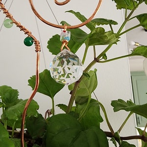 Close-up of a handmade plant stake suncatcher for garden decor, crafted with 16-gauge copper wire in a spiral design. The stake is adorned with colorful glass beads and features a millefiori glass bead hanging above a 20mm K9 crystal ball. The crystal sparkles in the sunlight, scattering rainbow light effects.