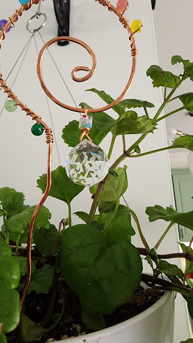 Close-up of a handmade plant stake suncatcher for garden decor, crafted with 16-gauge copper wire in a spiral design. The stake is adorned with colorful glass beads and features a millefiori glass bead hanging above a 20mm K9 crystal ball. The crystal sparkles in the sunlight, scattering rainbow light effects.