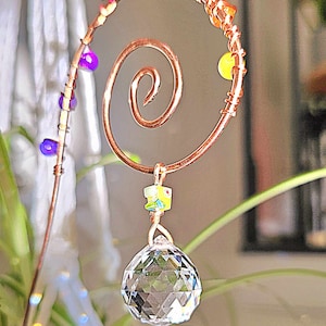 Close-up of a handmade plant stake suncatcher for garden decor, crafted with 16-gauge copper wire in a spiral design. The stake is adorned with colorful glass beads and features a millefiori glass bead hanging above a 20mm K9 crystal ball. The crystal sparkles in the sunlight, scattering rainbow light effects.